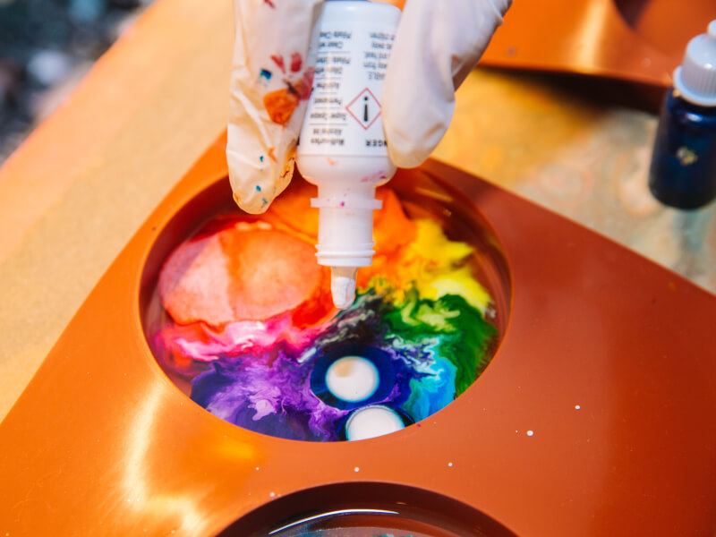 A close up of gloved fingers adding colours to a tray at a paint pouring class